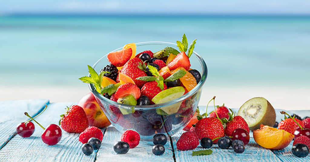 Cosa mangiare in estate - compertina - frutta di vari colori in una terrina di vetro e sparsa su un tavolo di legno in spiaggia